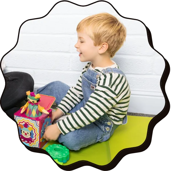 A young blonde boy wearing denim dungarees faces away from the camera holding a jack in the box toy