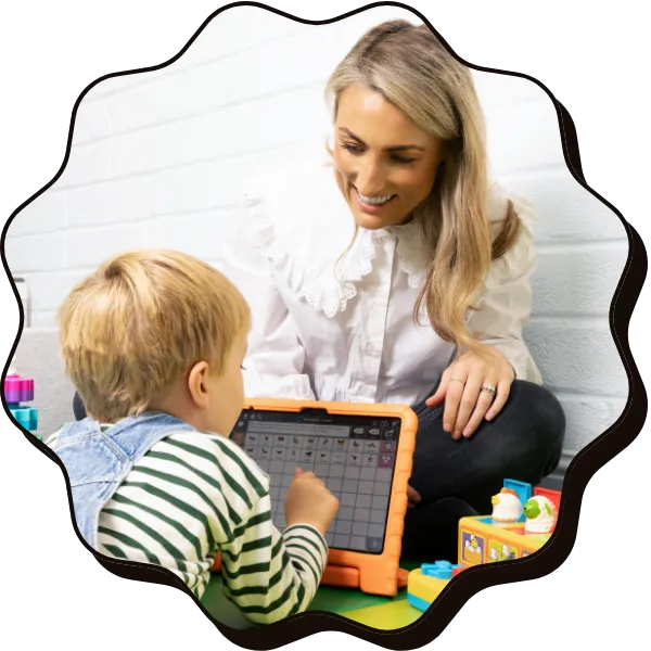 A blonde child faces away from the camera playing on a tablet, while a women with blond hair smiles at him, they are sitting on the floor
