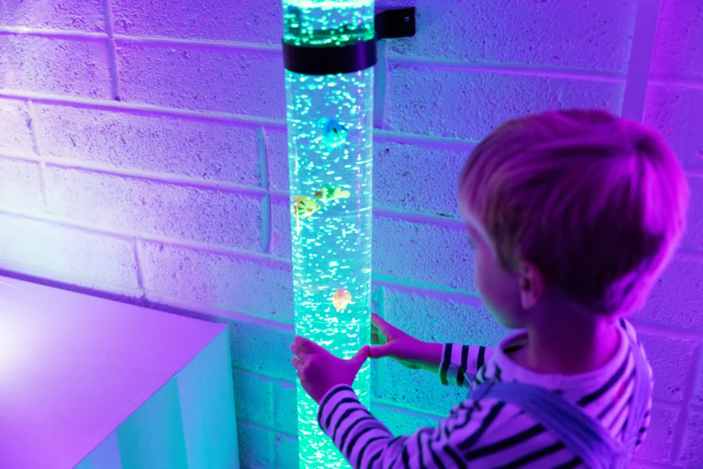 a small blonde boy faces away from the camera with his hands on a glowing light up fish sensory toy