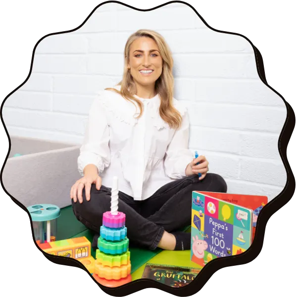 Julie, Senior Speech and Language Therapist, smiles at the camera. She is sitting on the floor surrounded by children's books and toys. She wears a white shirt with medium length blonde hair